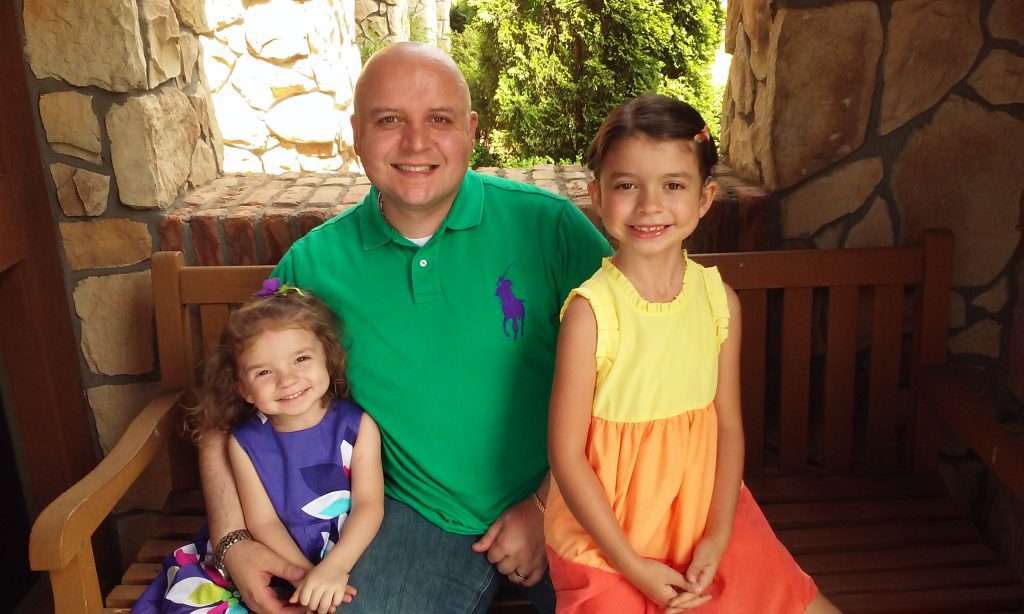 A grownup in a green polo shirt sitting on a wooden bench with two young girls in colorful dresses, smiling outdoors.