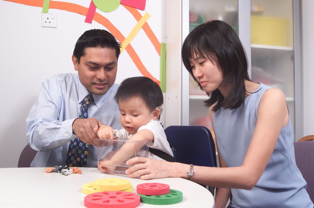A grownup and a woman assist a young child with a learning activity using colorful toys at a table.