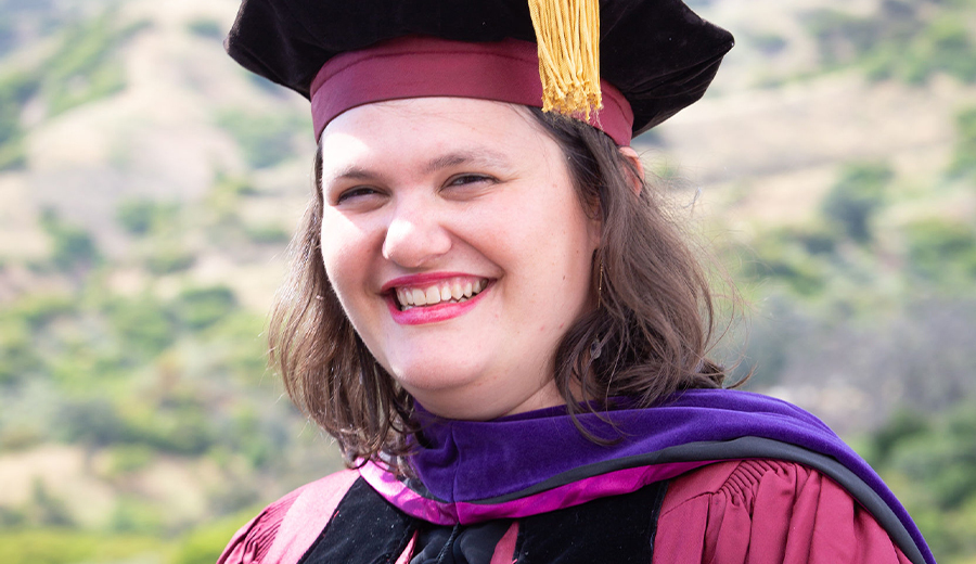 A joyful graduate with a disability in a purple and gold graduation cap and gown, smiling outdoors with a scenic, hilly background.