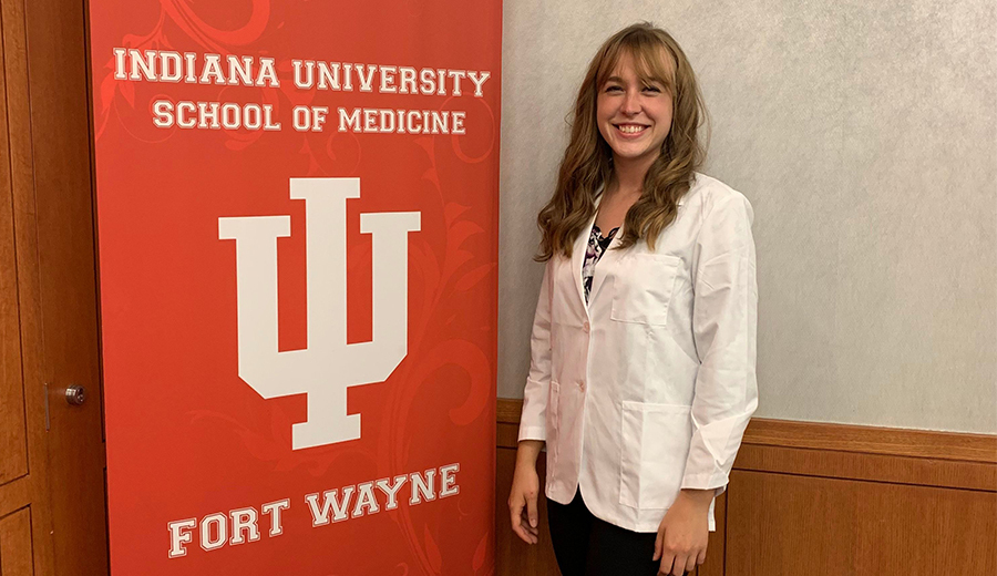 A smiling woman in a white lab coat stands next to a banner for Indiana University School of Medicine, Fort Wayne, accompanied by her child.