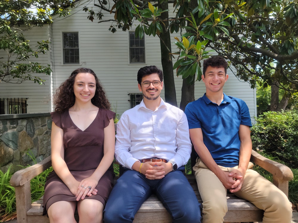 Three young adults smiling on a bench in front of a house with lush greenery, one using mobility aids due to a disability.