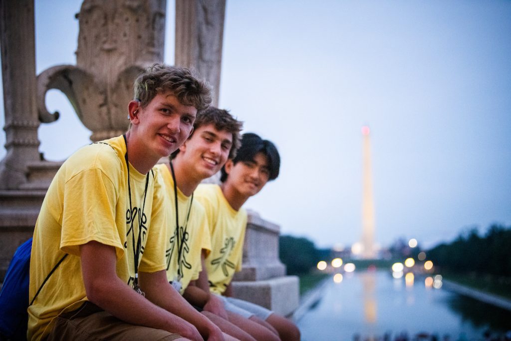 Three smiling young men, one using a wheelchair, sitting on a ledge by a waterway with the Washington Monument illuminated in the background at dusk.