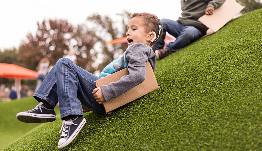 Child with a disability sliding down a grassy hill on a cardboard box with visible enjoyment, wearing casual clothing and sneakers.