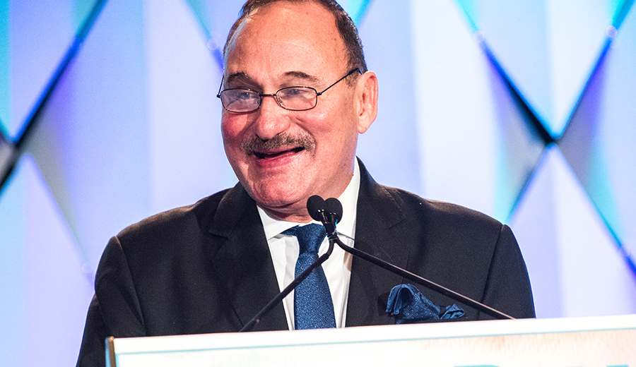 A man in a suit and glasses speaking at a podium during a family event, with a blue geometric background pattern.