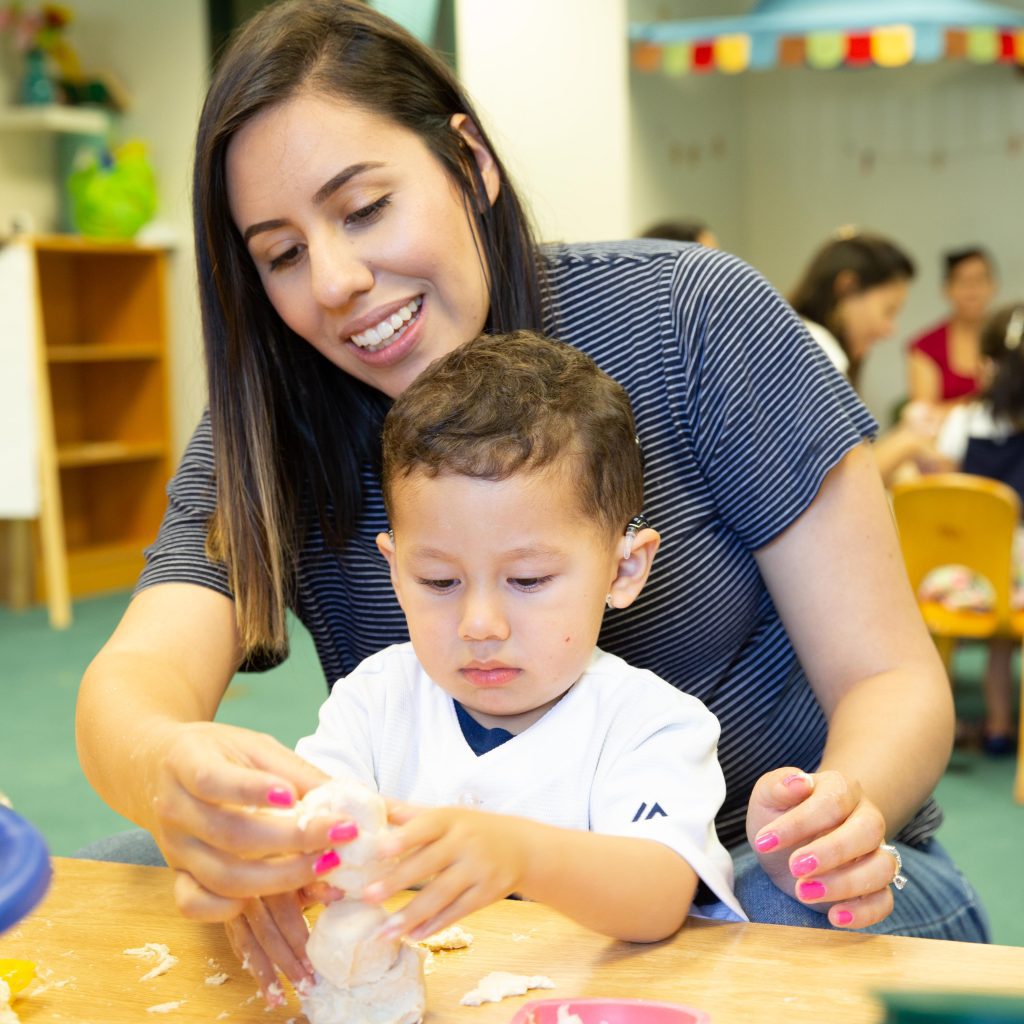 A grownup and a young boy engaging in a craft activity with clay at a table in a colorful classroom setting.