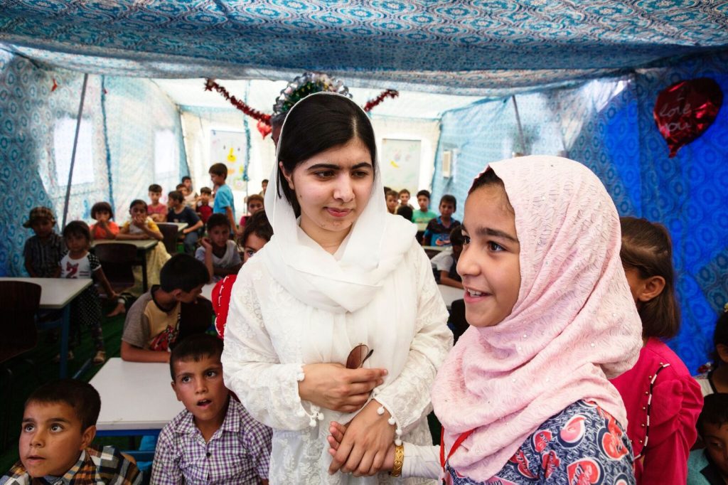 Malala Yousafzai holding hands with a young child in a classroom full of children, looking at each other and smiling.