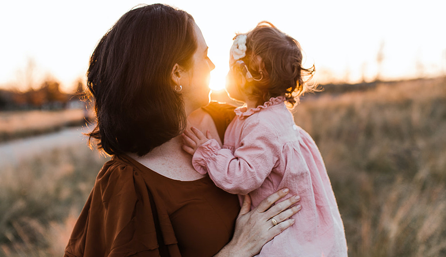 A mother holding her young daughter at sunset with warm light illuminating a natural field in the background, symbolizing family unity.