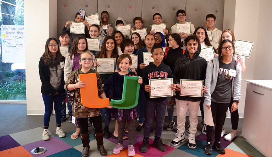 Group of diverse students proudly holding certificates and large letters inside a classroom with family assistance.