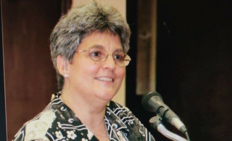 A grownup with short gray hair, wearing glasses and a floral shirt, speaking at a podium with a microphone.