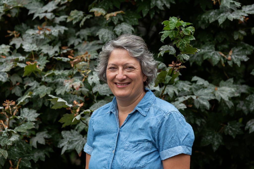 A smiling grey-haired woman in a blue denim shirt, using her hearing aid, stands in front of a green leafy background.