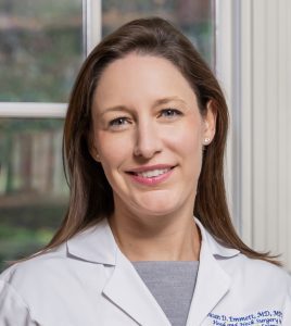 Portrait of a smiling grownup with long hair, wearing a white lab coat with an embroidered name and job title, standing against a window.