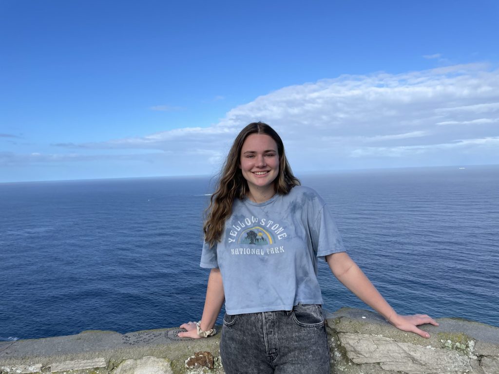 Young grownup in a t-shirt smiling on a coastal cliff with a clear blue sky and ocean in the background.