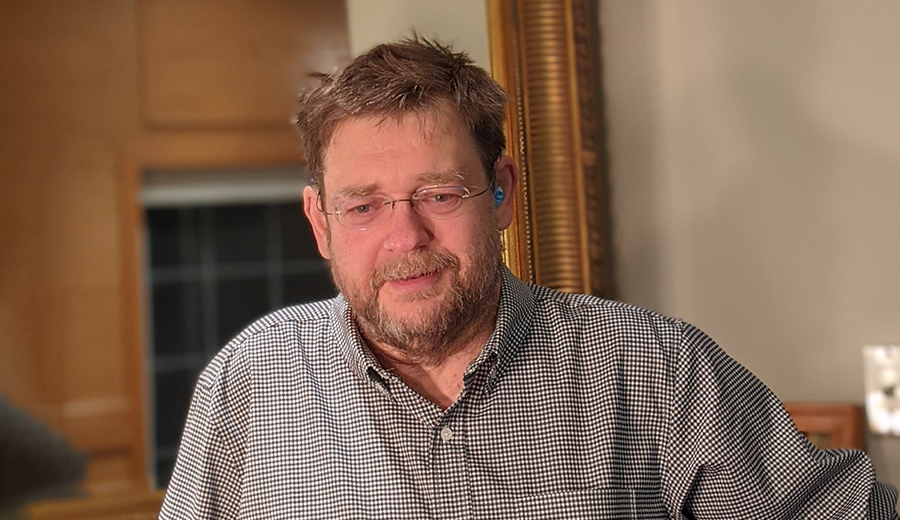 Middle-aged grownup with glasses and beard, wearing a checkered shirt, sitting indoors with a soft smile.