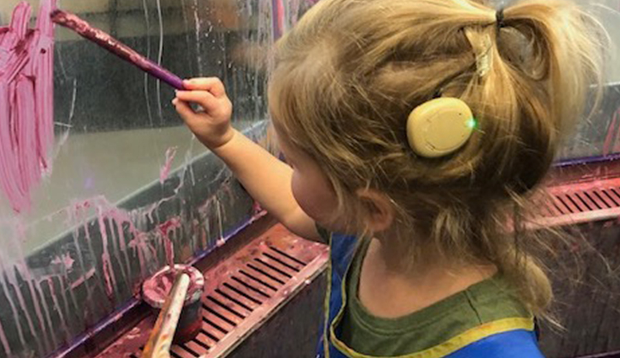 A young girl with a hearing disability paints on a glass surface with a pink brush.