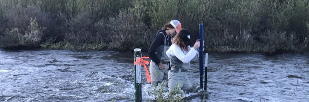 Two individuals utilizing surveying equipment beside a river, one grownup observing through the instrument while the other provides assistance.