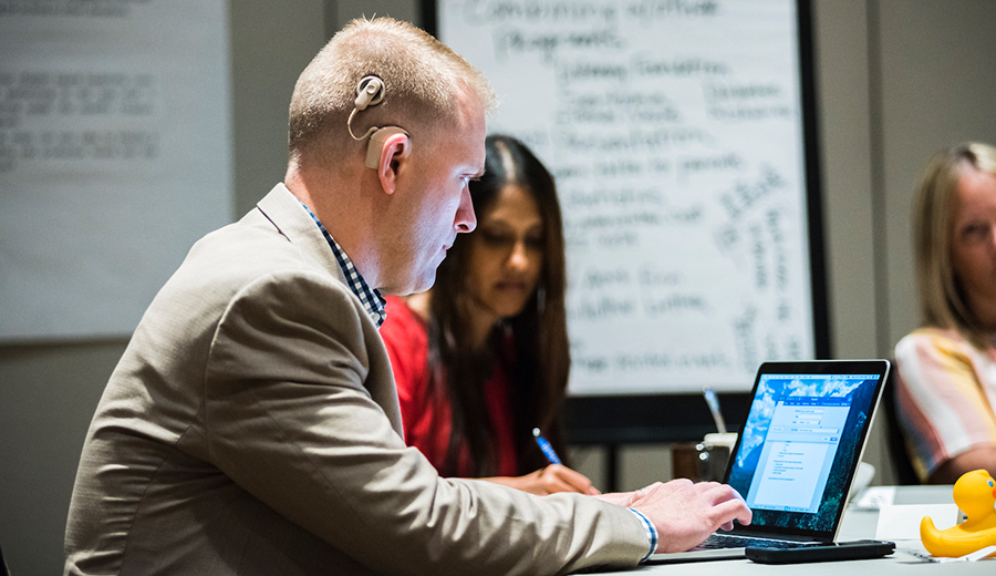 Man with a hearing aid using a laptop during a business meeting, requiring occasional assistance from colleagues, and a whiteboard in the background.