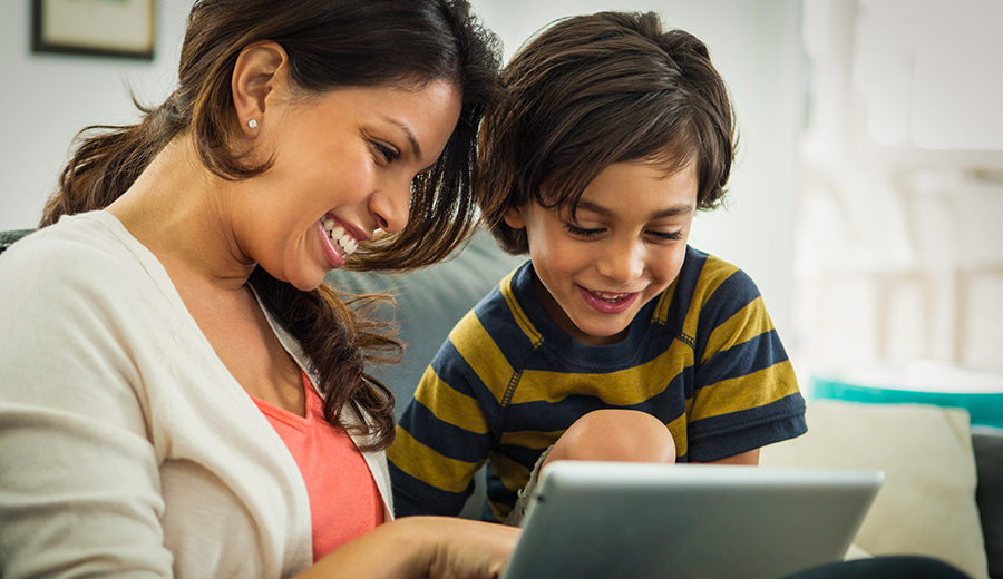 A woman and a young child smiling and looking at a tablet together while sitting on a couch.