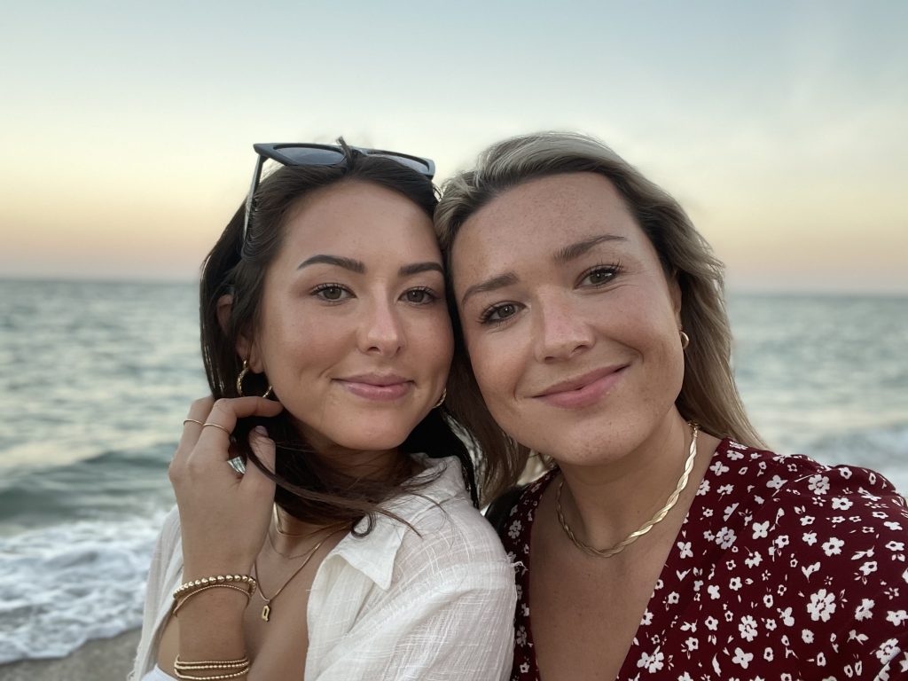 Two grownups taking a selfie on a beach at sunset, one with sunglasses on her head, both smiling at the camera.