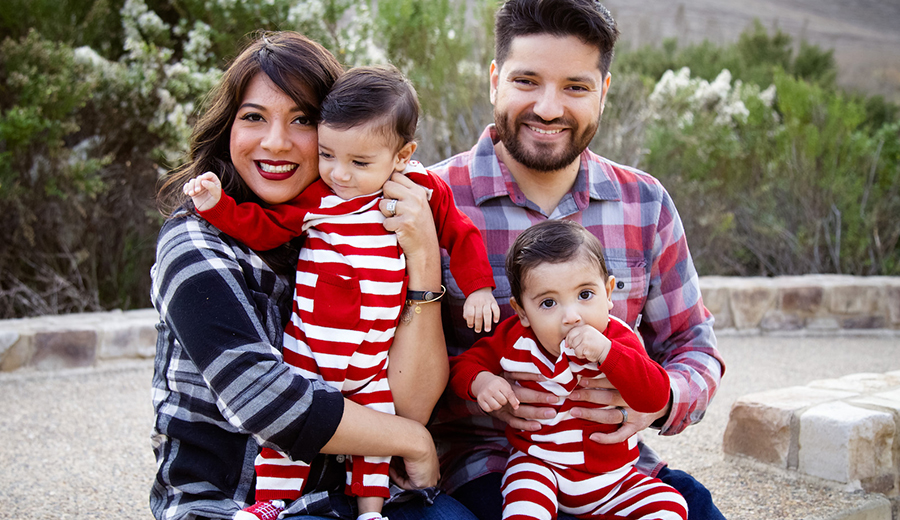 A family of four, with two adults and two young children, smiling and dressed in red and white striped outfits outdoors, offering assistance.