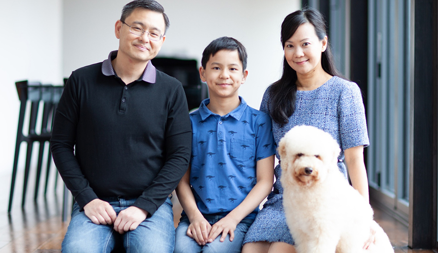 A family of three with their fluffy white dog posing together indoors, smiling at the camera as their child grins broadly.
