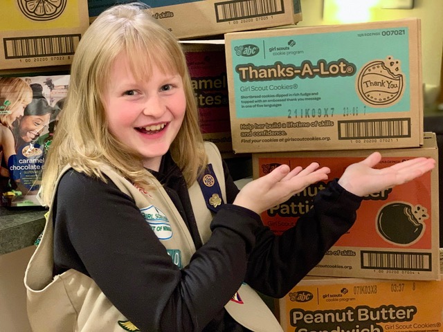 Young girl scout presenting boxes of cookies, expressing happiness, with a "thanks-a-lot" cookie box visible to her family.