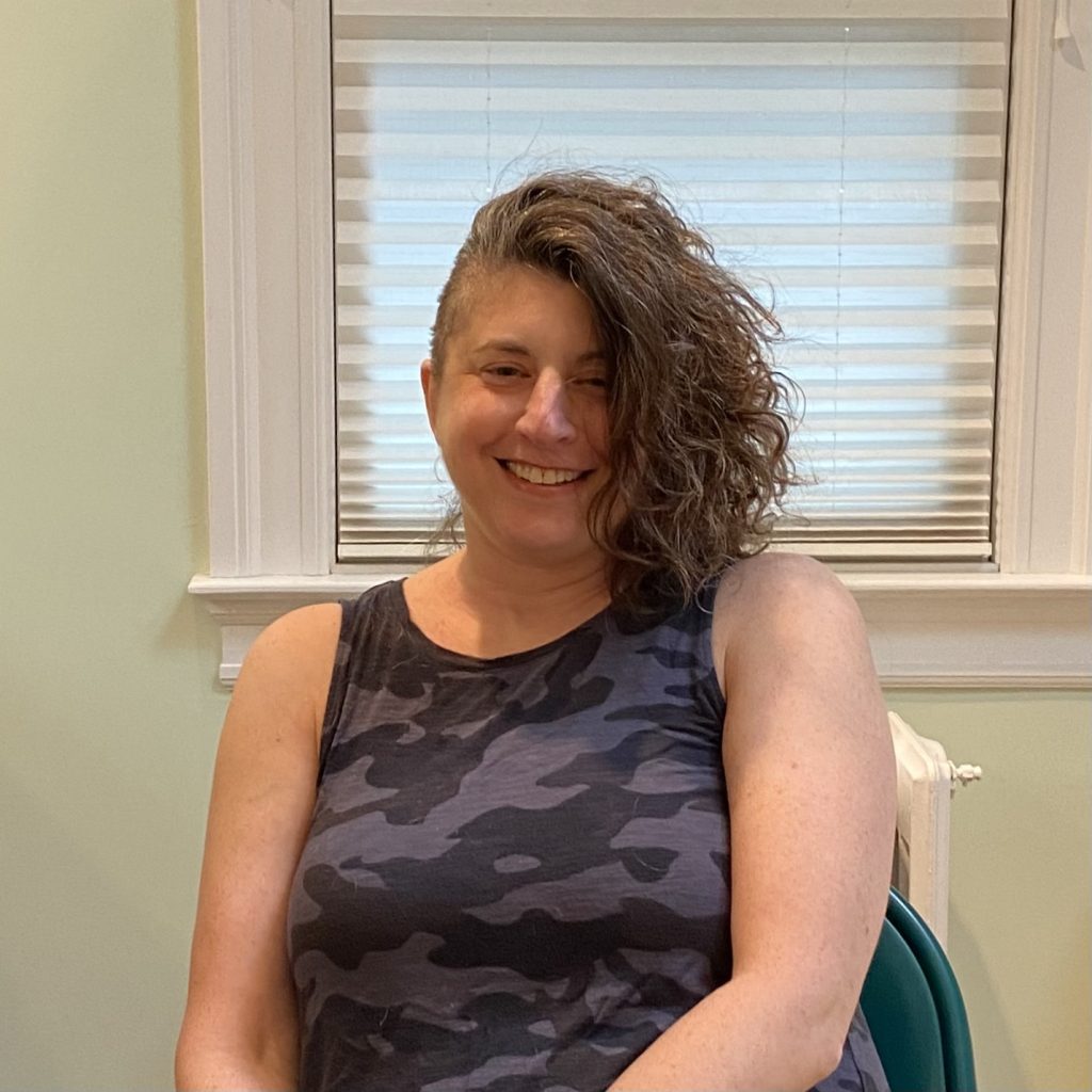 A woman with curly hair smiling while seated in a chair, wearing a camouflage tank top, with a window behind her.