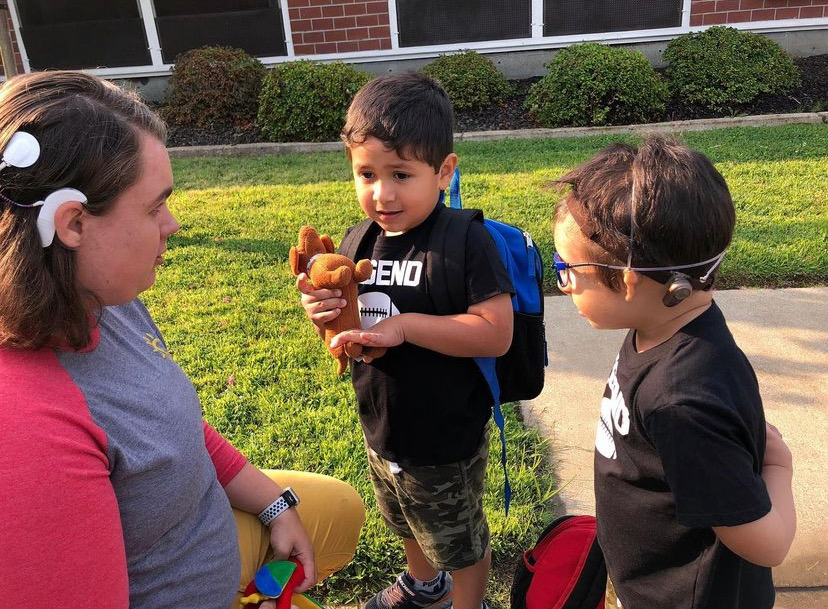 A woman wearing headphones kneels and offers assistance to two young boys, one with a soft toy and the other wearing sunglasses, outside a school building.