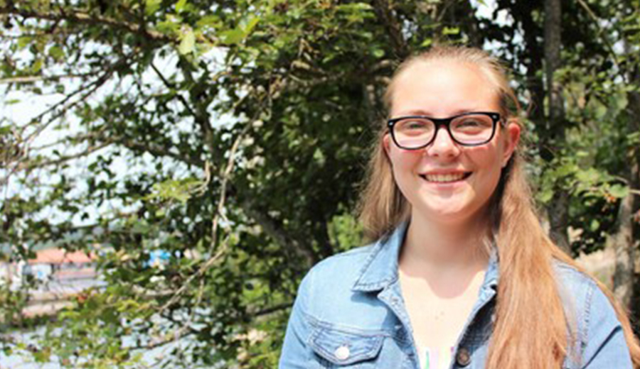 A young woman with glasses smiling, wearing a denim jacket, standing outdoors with trees and a blurry lakeside background.