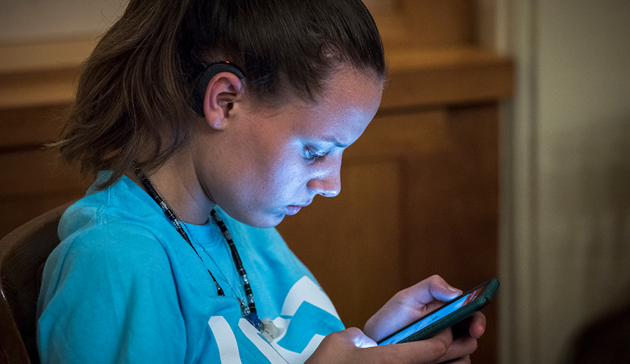 Young woman in a blue t-shirt focused on her smartphone as she hears her family's voices in the background, in a dimly lit room.