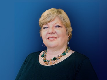 A portrait of a smiling middle-aged woman with short blond hair, wearing a green top and a colorful necklace, designed to hear family stories against a blue background.