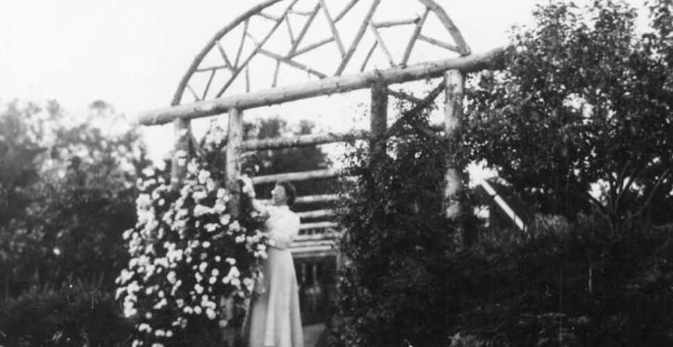 Two people in vintage attire, one with a disability, decorating a large dome-shaped trellis with flowers in a lush garden.