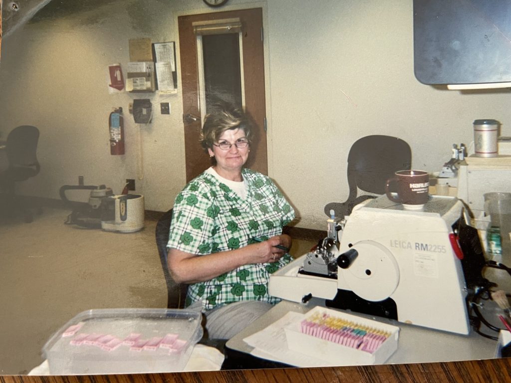 Woman in a checkered shirt using a microtome in a lab setting, with grownup assistance and lab equipment around.