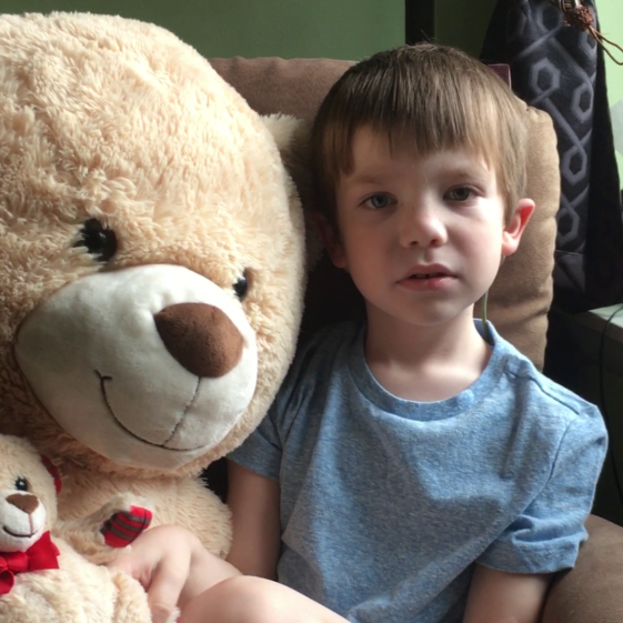 A young child with short hair sitting next to a large teddy bear on a sofa.