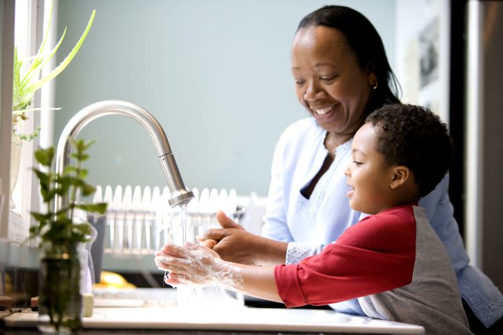 A woman and a young boy smiling while washing hands together at a kitchen sink, depicting a heartwarming family moment.