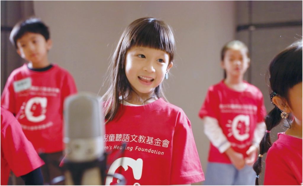 Young girl in a red shirt smiling near a microphone with other children in the background, one of whom has a disability.