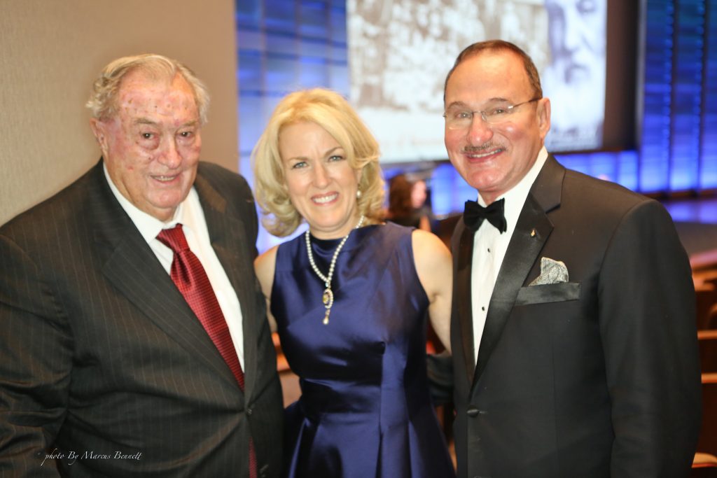 Two men in tuxedos and a woman in a blue dress smiling at a formal event, with a presentation screen visible in the background, discussing disability awareness.