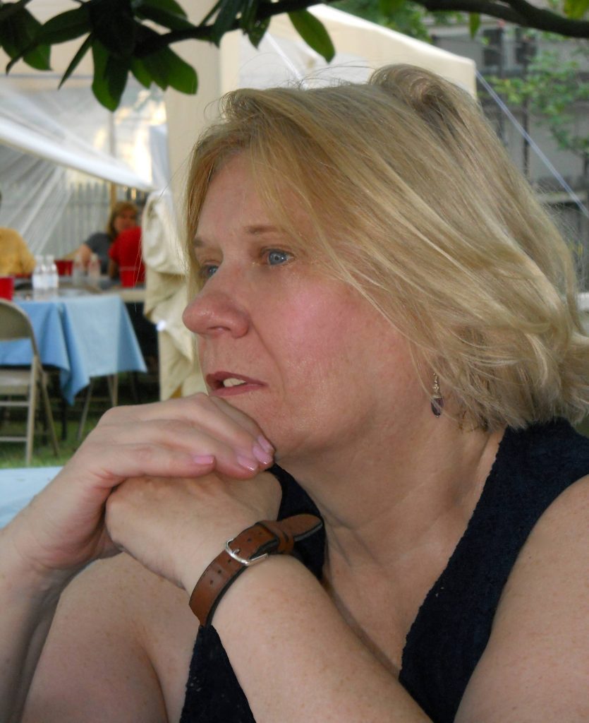 A thoughtful middle-aged woman with blonde hair, wearing a dark top and a wristwatch, resting her chin on her hand at a family outdoor event.