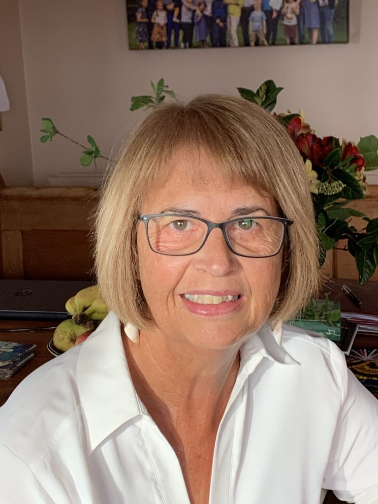 Smiling older woman with short blonde hair and glasses, wearing a white shirt, seated indoors with family paintings and fruit in the background.