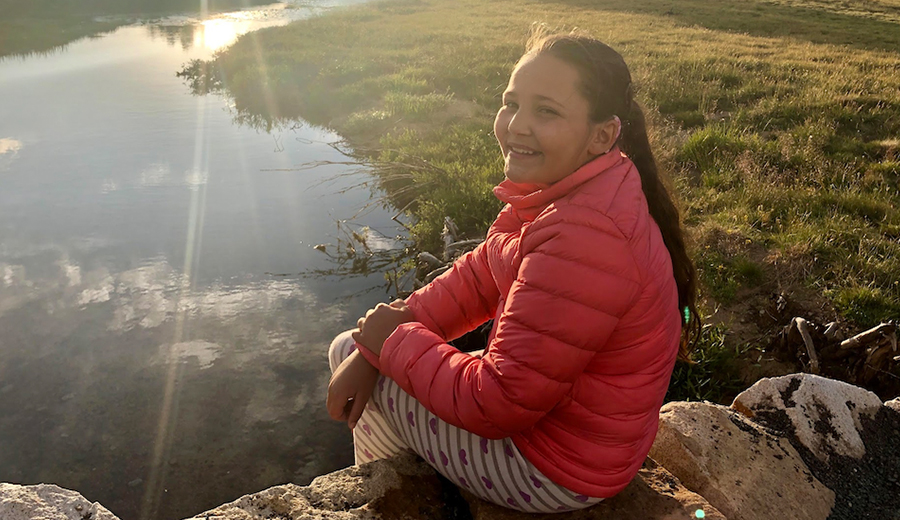 A young girl in a pink jacket smiling and sitting by a calm lake at sunset with her family.