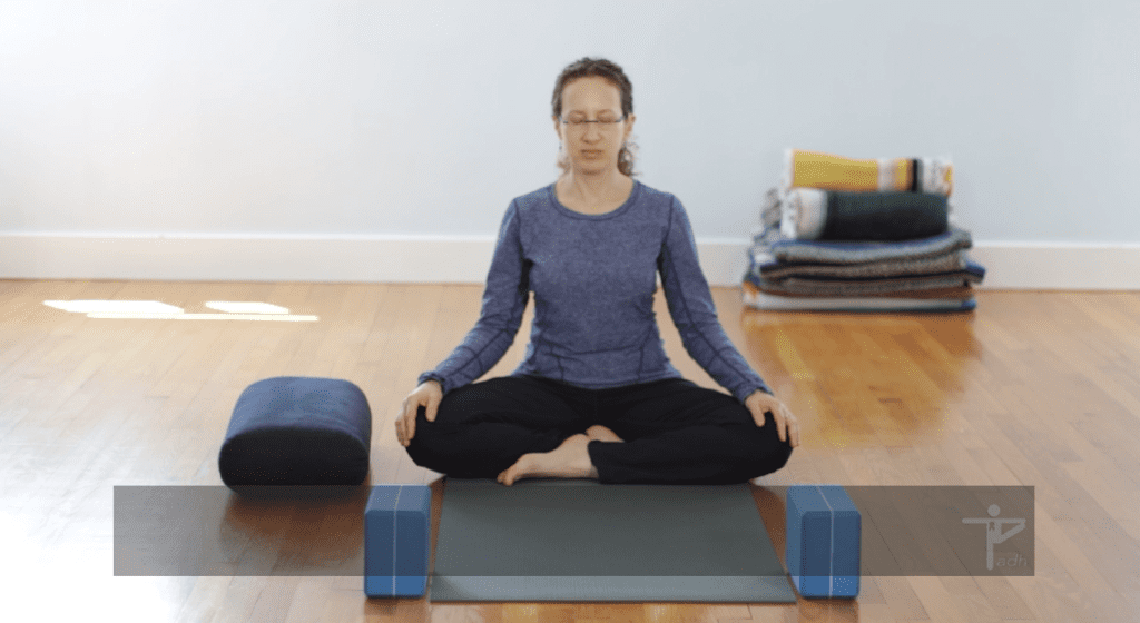 A grownup meditating in a cross-legged position on a yoga mat, with a bolster and blocks nearby in a serene room.