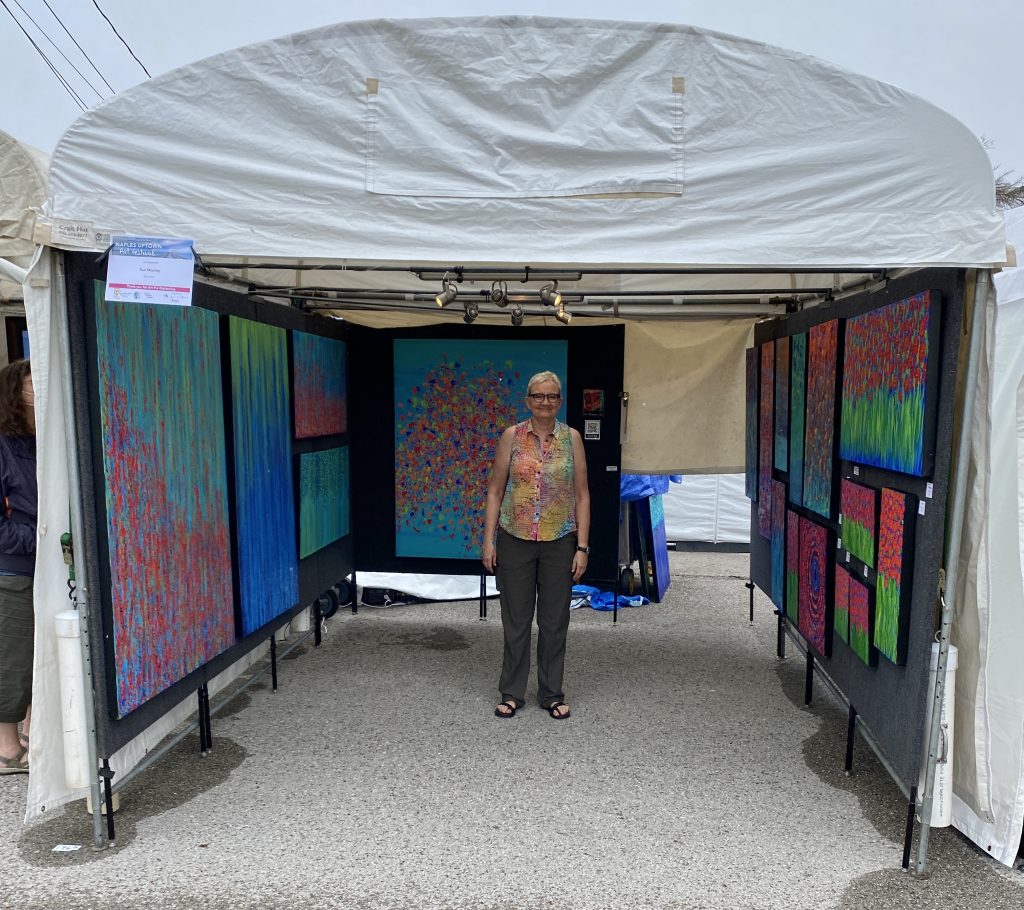 A woman stands in a white tent displaying colorful abstract paintings at an art fair, as her child explores nearby.