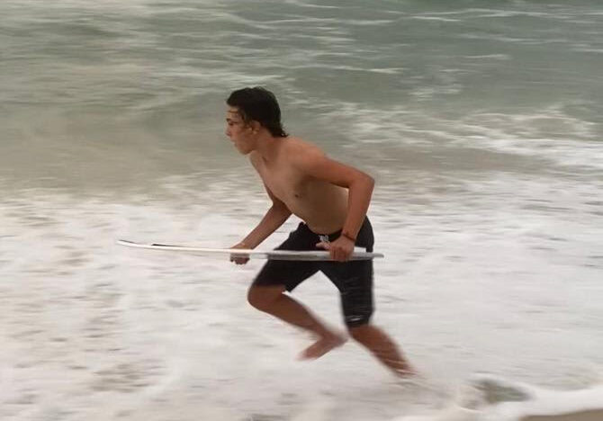 Young man with a hearing disability running along the beach with a surfboard under his arm, with a blurred motion background of the ocean waves.