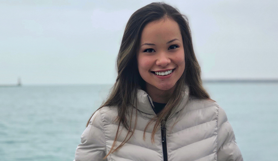 Young woman smiling, wearing a light gray jacket, with a calm sea and overcast sky in the background.