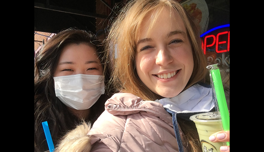 Two young women smiling for a selfie, one wearing a mask due to a disability, both holding bubble tea drinks outside a café.