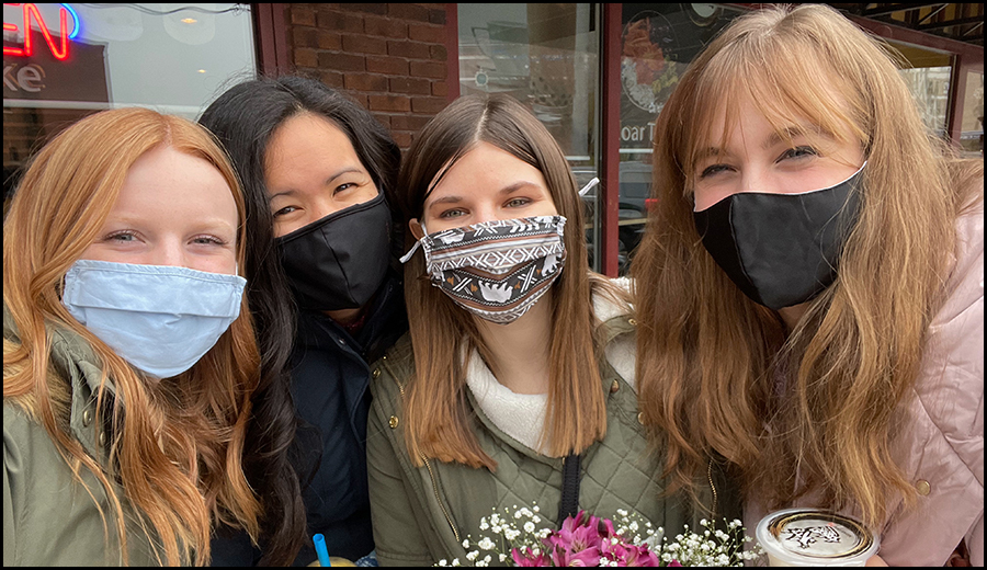 Four women with face masks smiling at the camera, holding flowers, standing in front of a brick building, offering assistance.
