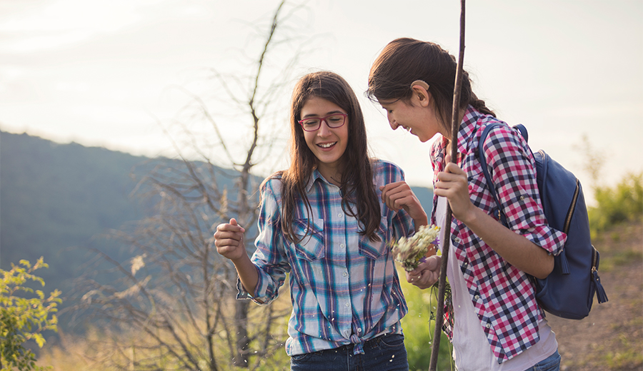 Two young women in plaid shirts and glasses walking outdoors, smiling and holding wildflowers, with a scenic family backdrop.