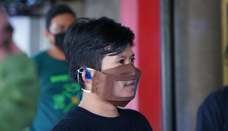 A person wearing a transparent face mask and a blue surgical mask underneath is looking to the side in a crowded area, trying to hear their family.