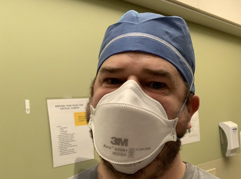 A man in surgical scrubs and a blue cap wearing an N95 mask indoors, with child-focused informational posters visible on the wall behind him.