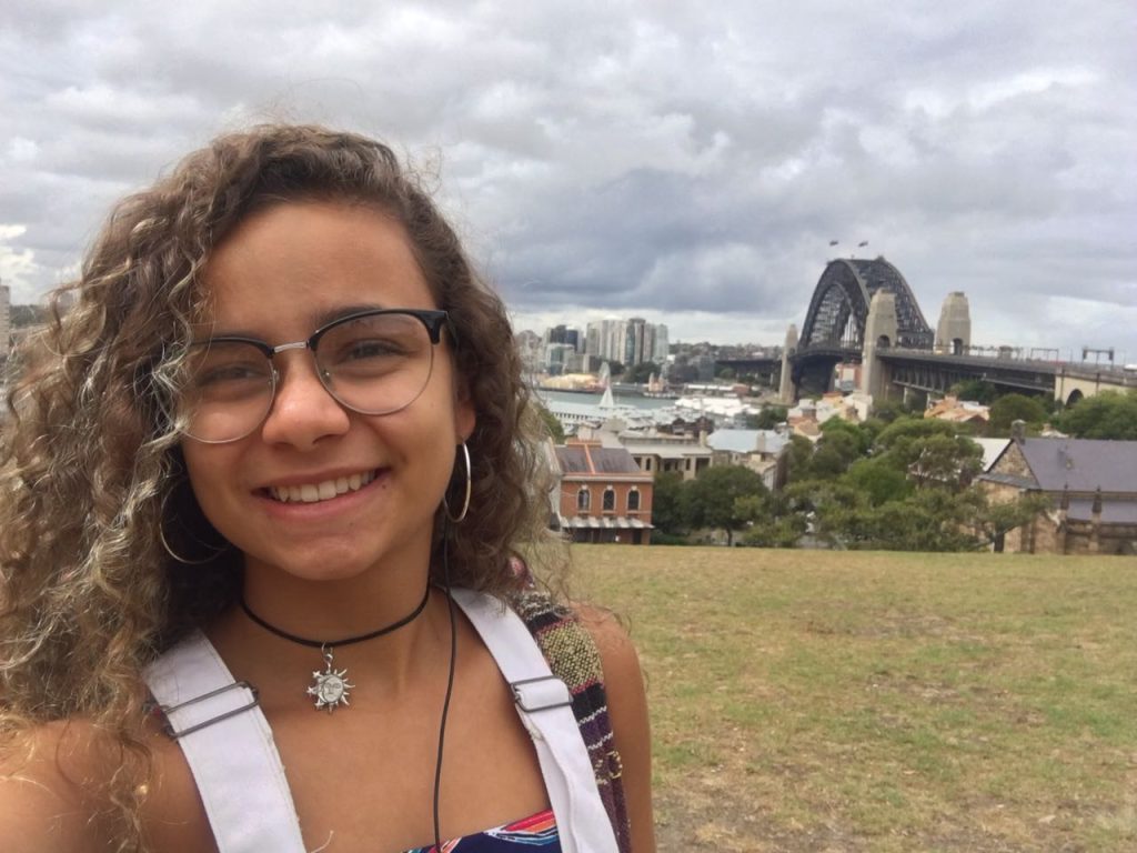 Young woman with curly hair smiling at the camera, with the Sydney Harbour Bridge in the background on a cloudy day.