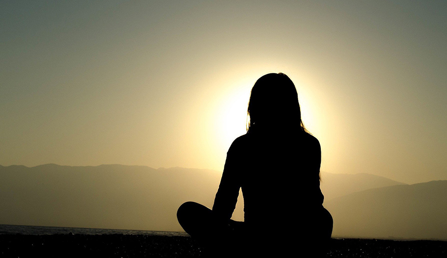 Silhouetted person sitting cross-legged on the ground against a sunset backdrop, straining to hear the distant call of nature.
